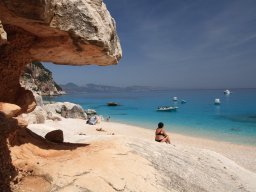Frau sitzt am Strand vor den Felsen von Cala Luna und schaut auf die Motorboote auf dem Meer Frau sitzt am Strand vor den Felsen von Cala Luna und schaut auf die Motorboote auf dem Meer